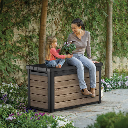Záhradný úložný box Keter DECO 380L Ashwood v záhrade, na ktorom sedia žena s dieťaťom v obklopení kvetín
