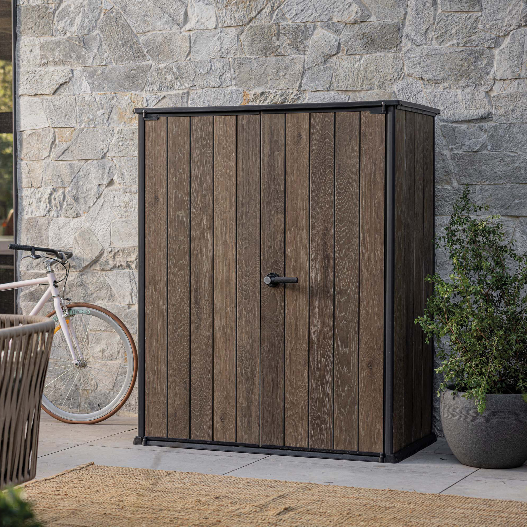 Záhradný domček Keter Deco Vertical 1400L Walnut s dvojitými dverami na terase pri kamennej stene