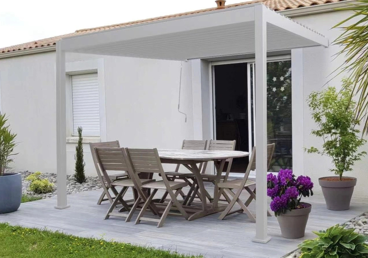 Modern&aacute; biela terasov&aacute; pergola so s&uacute;pravou dreven&eacute;ho z&aacute;hradn&eacute;ho n&aacute;bytku pri dome, obklopen&aacute; kvetin&aacute;čmi s rastlinami