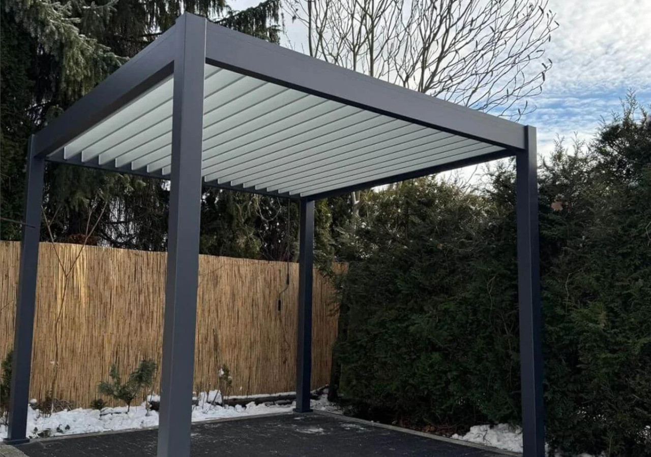 Antracitov&aacute; hlin&iacute;kov&aacute; pergola so svetl&yacute;mi lamelami na streche, umiestnen&aacute; na terase, na pozad&iacute; plotu z trstiny a zimnej veget&aacute;cie